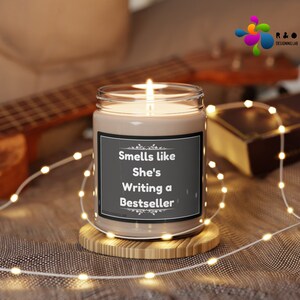 May include: A brown candle with a white label that reads "Smells like She's Writing a Bestseller". The candle is sitting on a wooden coaster and surrounded by white fairy lights.