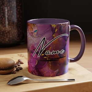 May include: A ceramic mug with a purple interior and handle, featuring a vibrant abstract design in shades of purple, gold, and blue. The word "Name" is written in a stylish script across the front. A spoon and a macaron are in the foreground.