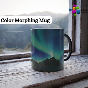 May include: A black color-changing mug with a picture of a green and blue aurora borealis over mountains. The mug is sitting on a wooden surface in front of a window.