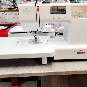 May include: White Bernina b38 sewing machine with a digital display and a variety of buttons. The machine is on a white table with a wood grain finish.