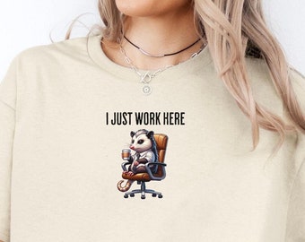 May include: A sand-colored t-shirt with a graphic of a possum sitting in a chair and the text "I just work here".