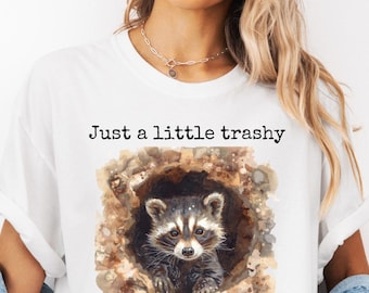 May include: White t-shirt featuring a watercolor illustration of a raccoon peeking out of a hole. The text "Just a little trashy" is printed above the raccoon. The shirt is a relaxed fit.