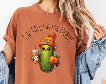 May include: A rust-colored t-shirt with the text "I'm falling for pickles" and a cartoon pickle wearing a hat and holding a pumpkin and a coffee cup.