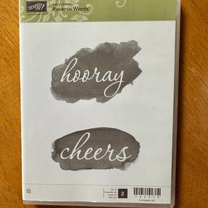 May include: Clear plastic case containing a stamp set. The set includes stamps with the words "hooray" and "cheers" in a handwritten style, over a watercolor-like background. The case has a green and white label with the text "Sale-A-Bration Reverse Words".