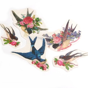 May include: A collection of five die-cut paper bird stickers. The birds are depicted in flight, with blue and brown plumage, and are adorned with floral bouquets of pink and purple roses. The stickers have a vintage, botanical aesthetic.