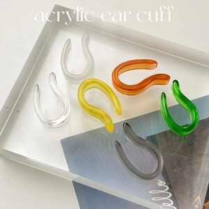 May include: Six acrylic ear cuffs in different colors: clear, yellow, orange, green, gray. The cuffs are shaped like a curved line.