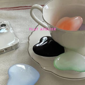 May include: A white teacup and saucer set with a silver trim, filled with heart-shaped stones in various colours. A clear phone case is visible in the background. The text "FLAT ATELIER" is printed on the image.