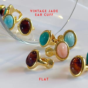 May include: An assortment of vintage jade ear cuffs in shades of turquoise, pink, and brown. The ear cuffs feature a gold-coloured metal setting and an oval-shaped stone. The image includes the text "VINTAGE JADE EAR CUFF".