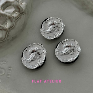 May include: Three silver-colored hair ties with a classical relief design. Each tie features a circular, ornate silver emblem with a detailed face and a black elastic band. The text "FLAT ATELIER" is visible in pink.