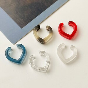 May include: Five acrylic ear cuffs in different colors and styles. The cuffs are shaped like a heart and are made of clear, translucent, and opaque acrylic.