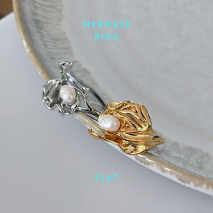 May include: Two rings, one silver and one gold, each featuring a pearl. The silver ring has a flowing, organic design, while the gold ring has a textured, layered appearance. The text "MERMAID RING" and "FLAT" are visible.