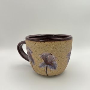 May include: A speckled tan ceramic mug featuring a dark brown rim and handle. The exterior is decorated with hand-painted light purple flowers with thin black stems. The mug has a rounded shape and a smooth, glossy finish on the rim and handle.
