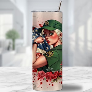 Puede incluir: Un vaso de acero inoxidable con una ilustración de dibujos animados de una mujer con uniforme y gorra verdes, flexionando su brazo frente a una bandera estadounidense. El diseño incluye rosas rojas y salpicaduras de sangre. Una pajita plateada está insertada en la tapa.