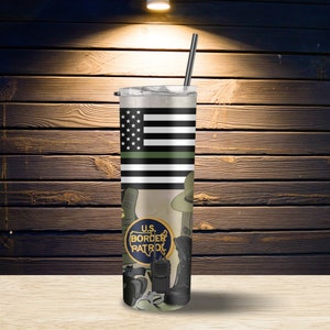 May include: A stainless steel tumbler with a green, white, and black American flag design. The tumbler features a green line, representing the thin green line, and the words "U.S. BORDER PATROL" in a circle. The tumbler is on a wooden surface.