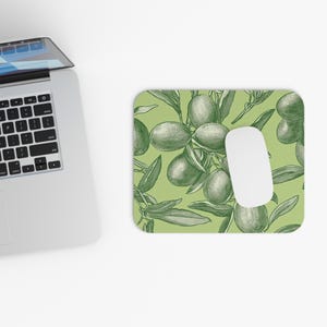 May include: A rectangular mousepad with a green background and a detailed olive branch design. The design features olives and leaves in a sketched style. A white computer mouse is on the mousepad, next to a laptop.