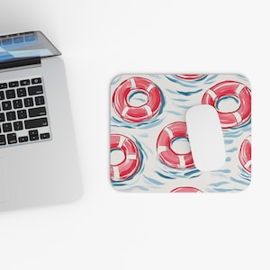 Puede incluir: Alfombrilla de ratón rectangular con un patrón de salvavidas rojos y blancos flotando sobre agua azul. Un ratón de ordenador blanco está posicionado sobre la alfombrilla. El diseño tiene un tema náutico, adecuado como accesorio de escritorio.