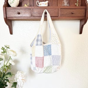 May include: A quilted patchwork tote bag featuring a variety of fabric squares in soft pastel colours including light blue, red, yellow, and white. The bag has a cream-coloured fabric trim along the edges and two integrated shoulder straps.