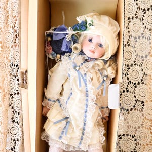 May include: A porcelain doll with blonde curls, blue eyes, and a cream-coloured bonnet. The doll wears a cream dress with blue ribbon accents, lace trim, and white lace tights. It is wearing white lace-up boots and is packaged in a cardboard box.