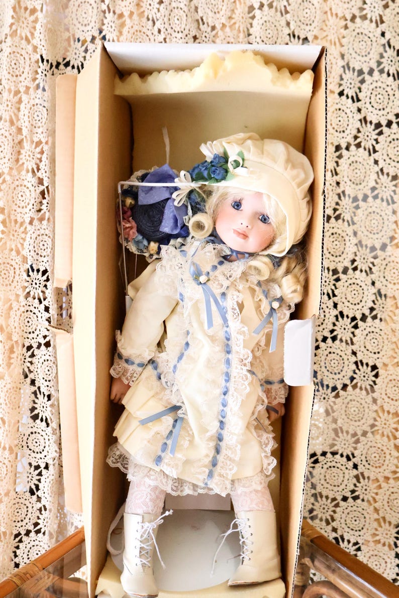 May include: A porcelain doll with blonde curls, blue eyes, and a cream-coloured bonnet. The doll wears a cream dress with blue ribbon accents, lace trim, and white lace tights. It is wearing white lace-up boots and is packaged in a cardboard box.