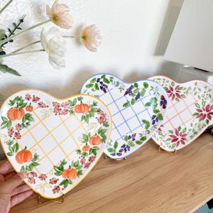 Peut inclure: Trois assiettes en céramique en forme de cœur avec des motifs décoratifs. L'une présente des citrouilles oranges et du feuillage d'automne sur une grille jaune. Une autre a des baies bleues et une grille bleue. La troisième a des fleurs rouges et une grille rose.