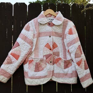 Puede incluir: Una chaqueta acolchada con diseño de patchwork en tonos blanco y rosa empolvado. La prenda incluye un cuello redondeado, mangas largas, dos bolsillos delanteros y un cierre frontal de botones con patrones circulares en forma de abanico.