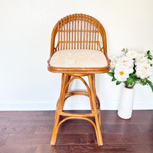 May include: A natural rattan bar stool with a curved back and a patterned cream-colored seat. The stool has a swivel design and a sturdy base. The overall color is a warm, light brown. The stool is approximately 40 inches tall.