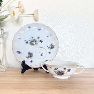 May include: A white teacup and saucer set with a floral violet design. The scalloped-edge saucer and teacup are adorned with purple violets and gold trim. The teacup has two handles and a matching violet pattern.