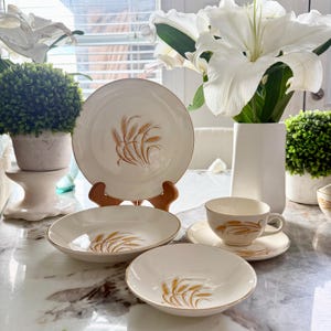 May include: A cream-colored vintage dinnerware set featuring a wheat stalk design in gold. The set includes a large plate, bowls, a teacup, and a saucer. The rims of the plates and teacup are trimmed with gold.