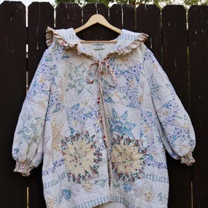 May include: A long-sleeved quilted jacket featuring a patchwork star pattern in shades of blue, yellow, and white. The garment includes a ruffled collar, elasticized cuffs, and a front tie closure, all constructed from various floral and striped fabrics.
