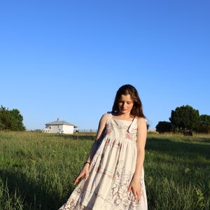 May include: A cream-colored sundress with floral and lace details. The dress has a square neckline and spaghetti straps. The dress is worn outdoors in a field of tall green grass under a clear blue sky.