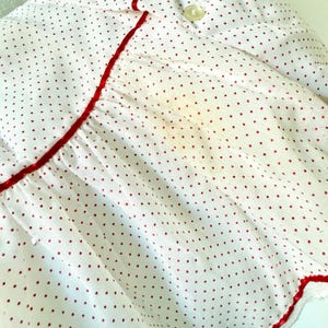 May include: White fabric with a red polka dot pattern. The garment has red trim along the edges and a button closure. The fabric is slightly wrinkled, and there is a small stain.