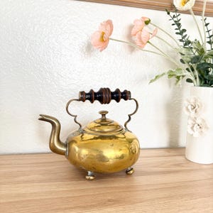 May include: A vintage brass teapot with a dark wooden handle and curved spout. The teapot has a round body and sits on three small feet. The handle is attached to the body with decorative brass supports. The teapot is displayed on a wooden surface.