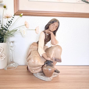 Puede incluir: Figura de cerámica de una mujer sentada, apoyando la cabeza en la mano. Viste un vestido largo y sostiene una pequeña jarra. La figura está pintada en tonos apagados de marrón, beige y crema, con un aspecto envejecido.