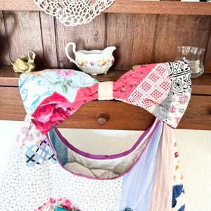 May include: A large fabric bow featuring a patchwork design with various patterns including floral prints, red and white gingham, and black and white geometric motifs. The bow is held together by a solid cream-coloured centre band made of fabric.