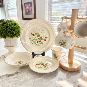 Può includere: Un set di piatti color crema con un design a fragole. Il set include piatti, ciotole e tazze, con un albero di tazze in legno. I piatti e le ciotole hanno un bordo dorato. È visibile anche una piccola pianta verde in un vaso bianco.