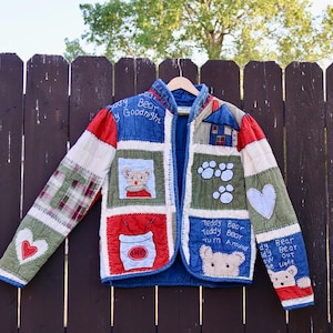 Könnte beinhalten: Eine gesteppte Jacke mit Patchwork-Design aus verschiedenen Stoffquadraten. Die Jacke zeigt gestickte Teddybären-Motive, Herzen, Pfotenabdrücke und den Text "Teddy Bear Teddy Bear Turn Around" sowie "Teddy Bear Teddy Bear Out the Light."