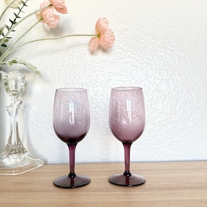 May include: Two matching purple wine glasses with textured glass. The stems and bases are a darker shade of purple. The glasses are displayed on a wooden surface with a white wall in the background. Pink flowers are in the background.