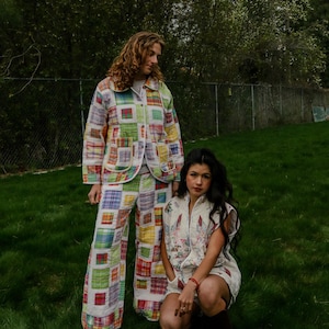May include: Two people wearing patchwork quilted outfits. One person stands in a matching jacket and wide-leg pants. The other person is kneeling in a quilted vest and shorts. The outfits feature a mix of colorful squares.