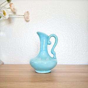 May include: A light blue ceramic jug with a curved handle and a rounded base. The jug is approximately 20 cm tall and sits on a wooden surface. The background is a white wall with a few flowers.