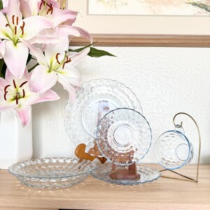 Puede incluir: Un conjunto de vajilla de vidrio transparente con un patrón texturizado en forma de burbujas. El juego incluye platos, platillos y una taza de té con su platillo a juego. Un jarrón con lirios rosas se encuentra en el fondo.
