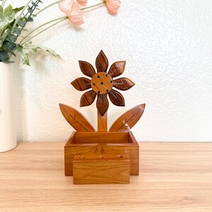 May include: A wooden sunflower-shaped desk organizer. The flower has dark brown petals and a lighter brown center. The leaves and box are light brown. The organizer includes a rectangular box and a smaller compartment, perfect for pens and pencils.