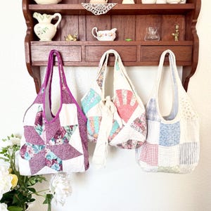 May include: Three handmade quilted tote bags hanging on a wooden rack. The left bag features a purple floral star pattern. The centre bag has a fan-shaped design with cream ties. The right bag displays a blue, pink, and white checkered patchwork.