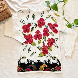 May include: Cream-colored sweater with a floral design featuring red flowers and green leaves. The bottom hem has a black border with paisley and geometric patterns. The long-sleeved sweater is made of a knitted material.