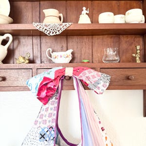 May include: A patchwork tote bag featuring a large fabric bow at the top, constructed from various textile scraps in shades of pink, blue, white, and black with floral, plaid, and striped patterns, hanging from a wooden shelf with kitchen decor.