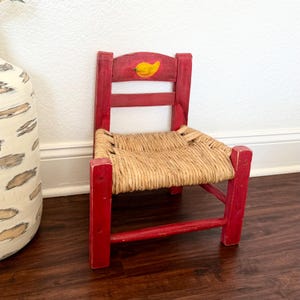 Op de afbeelding: Een kleine, rode houten stoel met een geweven strooien zitting. De rugleuning van de stoel heeft een geel chilipeperontwerp. De stoel is rood geschilderd en heeft een rustieke, handgemaakte uitstraling. De stoel is geschikt voor een kind.