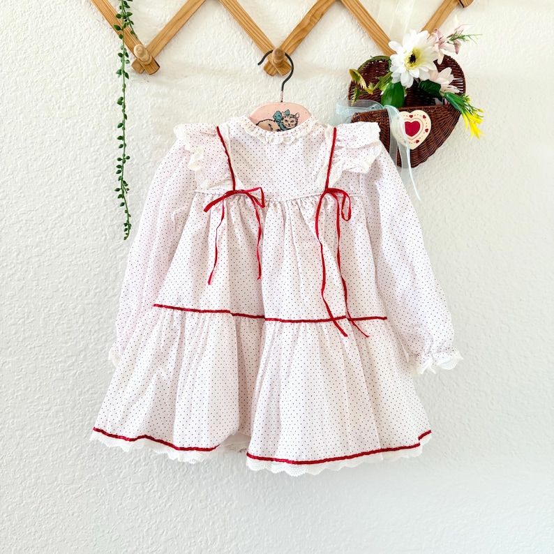 May include: A white and red polka dot dress with long sleeves, red trim, and red ribbon ties. The dress has a tiered skirt and ruffled details. It is hanging on a wooden hanger.