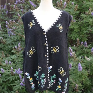 May include: A black sleeveless knit vest featuring embroidered yellow and black bees scattered across the front, with a floral pattern along the bottom hem. The garment has a V-neckline trimmed with a black and white checkered border and buttons.