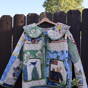 May include: A quilted jacket featuring a patchwork design with various printed motifs including surfboards, a yellow car, a blue bird, and a squirrel. The jacket has a large collar, long sleeves, two front pockets, and a tie-front closure with green fabric.