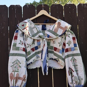 May include: A cropped long-sleeve jacket made from quilted fabric featuring a patchwork design of floral patterns, birdhouses, and geometric shapes in shades of red, blue, green, and white. The jacket has a wide collar and a front tie closure.