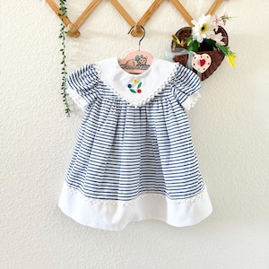 以下が含まれることがあります： 白と青のストライプ柄のワンピース。白い襟と裾が特徴です。襟には刺繍の花が飾られています。ワンピースは半袖で、木製のハンガーに掛けられています。